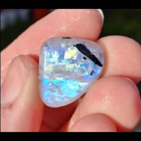 Light toned hand holding rainbow colored triangular shaped rainbow moonstone with a white background. 