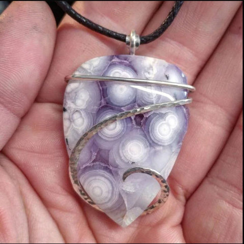 Purple with white spots luna agate stone wrapped in a band of sterling silver pendant on a black string being held by a human hand