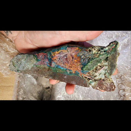 Persons hand holding a multicolored piece of chalcedony colored with copper metal and copper oxides. 