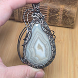 A hand holds a teardrop-shaped agate stone pendant wrapped in silver-toned wire, with a carved wooden box blurred in the background.