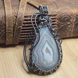 A teardrop-shaped pendant featuring a gray and white banded agate stone, wrapped in dark silver-toned metal with decorative wire curls and a small bow at the top, displayed against a rustic brown leather background.