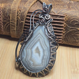 A handmade teardrop pendant featuring a gray and white banded agate stone, wrapped in dark silver wire with gold-toned accents at the bottom, displayed on a wooden surface in front of a brown leather strap.
