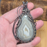 A hand holding a teardrop-shaped agate stone pendant wrapped in dark silver wire, with a copper-toned accent at the top and along the lower edge, set against a wooden background.