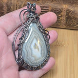 A hand holding a teardrop-shaped agate stone pendant wrapped in dark silver and copper wire, with a rustic wooden background.