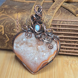 A heart-shaped druzy gemstone pendant with a copper wire wrap and a small light blue stone accent, displayed on a wooden surface with a leather strap in the background.