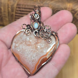 A heart-shaped agate druzy pendant with copper wire wrapping and a small light blue stone, held in a person's hand.