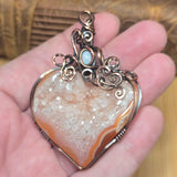 A heart-shaped druzy stone pendant with copper wire wrapping and a small light blue stone accent, held in a person's hand against a warm wooden background.