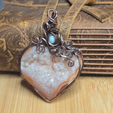 A heart-shaped druzy agate pendant with swirling copper wire, featuring a small light blue stone accent at the top, displayed on a wooden surface in front of a leather strap.