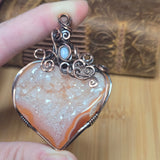 A heart-shaped druzy gemstone pendant with a copper wire wrap and a small light blue stone accent, held by a hand over a wooden background.