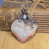 A heart-shaped druzy stone pendant wrapped in copper and silver wire, featuring a small light blue stone accent at the top, displayed on a wooden surface in front of a rustic brown leather book.