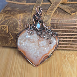A heart-shaped druzy agate pendant with a copper wire wrap and a small light blue stone, displayed on a wooden surface in front of a brown leather strap.