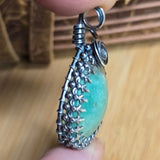 Green stone pendant with silver decorative frame held by a hand on a wooden surface