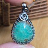 Teardrop-shaped pendant with green stone and silver spiral design held between fingers.