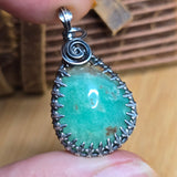 Teardrop-shaped pendant with green stone and silver wire wrapping held between fingers.