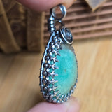Teal stone pendant with silver filigree design held by a hand on a wooden surface