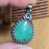 Teardrop-shaped pendant with green stone and silver frame held between fingers on a wooden surface.
