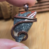 Close-up of a copper-wrapped stone pendant held between fingers on a wooden surface.