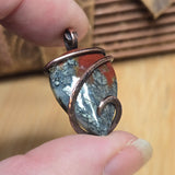 Close-up of a stone pendant with copper wire wrapping held between fingers on a wooden surface.