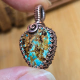 Turquoise stone pendant with copper wire wrapping held between fingers against a blurred background