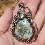 Ornate copper pendant with a central gemstone held in a hand against a blurred background