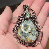 Copper wire-wrapped pendant with a natural stone center held in a hand.