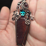 Red moss agate coffin pendant with antiqued copper wirework, garnet accents and an iridescent green skull.
