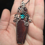Red moss agate coffin pendant with antiqued copper wirework, garnet accents and an iridescent green skull.