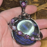 Close-up of a hand holding a copper pendant with purple gemstones against a textured background.