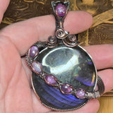 Hand holding a copper pendant with a large gemstone and smaller purple stones on a textured background