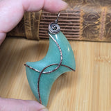 A handmade abstract form pendant made of polished light green stone with copper wire wrapping, held by a hand over a wooden surface.