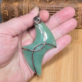 A handmade pendant made of polished light green aventurine with copper wire wrapping, held in a person's hand against a bamboo background.