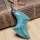 A handmade pendant made from a polished aventurine crescent moon-shaped stone wrapped with textured silver wire, resting on a rustic brown leather strap and wooden surface.