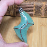 A hand holds a crescent-shaped aventurine pendant with textured copper wire details and a copper spiral top, set against a wooden background.