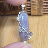 Hand holding a purple crystal pendant with silver wire wrapping on a wooden surface