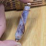 Hand holding a purple stone pendant with copper wire wrapping on a wooden surface
