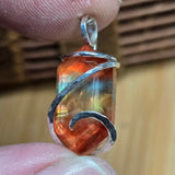Multicolored Ammolite pendant with silver wire wrapping held between fingers against a wooden background