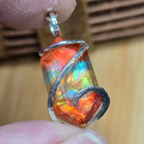 Multicolored Ammolite pendant with silver wire wrapping held between fingers against a wooden background