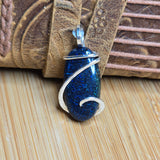 A blue glitter pendant with a silver wire-wrapped design, resting on a light wood surface in front of a brown leather strap.