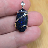 A close-up of a hand holding a small oval pendant with a dark blue glittery stone wrapped in silver-colored metal.