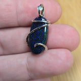 A close-up of a hand holding a small oval pendant with a dark blue glittery stone and silver wire wrapping, set against a light wood background.