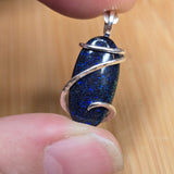 A close-up of a hand holding a small oval pendant with a dark blue glittery center and silver wire wrapping, set against a light wood background.