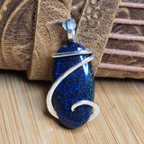 A blue glitter pendant with a silver wrap design, resting on a textured brown leather strap over a light wood surface.