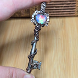 Silver key-shaped object with a colorful gemstone held between fingers against a wooden background