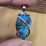 Blue stone pendant with copper wire wrapping held between fingers on a wooden surface