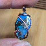 Blue pendant with copper wire wrapping held between fingers against a wooden background