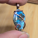 Opal pendant with copper wire wrapping held between fingers against a wooden background