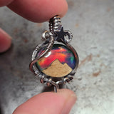 Jewelry with a colorful stone held between fingers against a neutral background