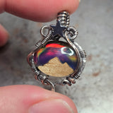 Silver pendant with a colorful stone held between fingers against a neutral background