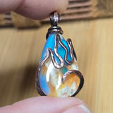 Teardrop-shaped pendant with flame design held between fingers against a wooden background