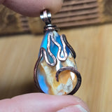 Teardrop-shaped pendant with flame design held between fingers against a wooden background